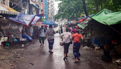 People walking through an outdoor market in the rain, some are holding umbrellas. The market stalls are covered by awnings and tarpaulin.