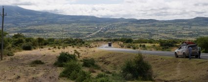 Car parked at the top of a winding road, with herds of cows are walking beside the road. There is a large expanse of open land in the distance, with fields and trees surrounded by mountains.